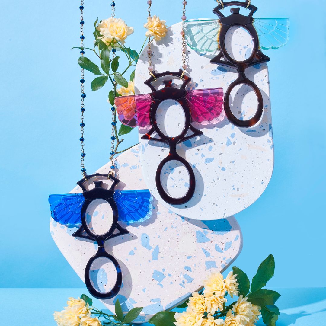 Colgante-gafas en forma de escarabajo egipcio. Realizado en combinación de acetato en tres opciones, azul, rosa y verde. Una bisagra hace que se pueda transformar en gafas de lectura