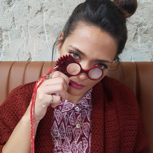 Mujer utilizando Colgante-medallón gafas en forma de clavel. Realizado en acetato de varias capas de color rojo, con abalorios y cordón en rojo con remates y fornituras plateados. Una bisagra hace que se pueda transformar en gafas de lectura.