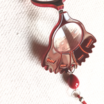 Colgante-medallón gafas en forma de clavel. Realizado en acetato de varias capas de color rojo, con abalorios y cordón en rojo con remates y fornituras plateados. Una bisagra hace que se pueda transformar en gafas de lectura.